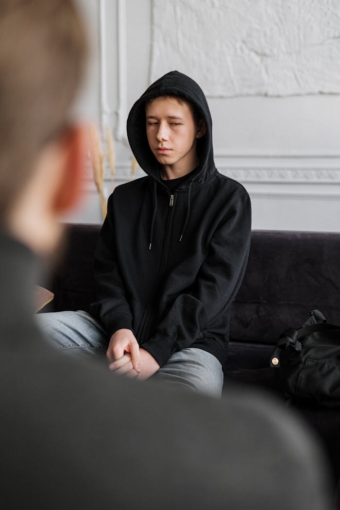 A teenager in a black hoodie engages in a counseling session, sitting calmly indoors.